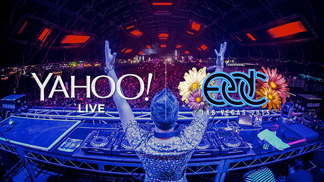 A DJ standing behind a DJ booth during a festival with the sponsor Yahoo! and EDC Las Vegas written on top of him.