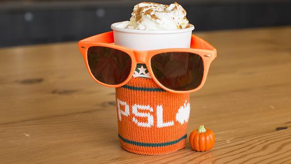 A to-go coffee cup from Starbucks with a PSL knit sleeve, a pair of orange sunglasses, and a small plastic pumpkin.